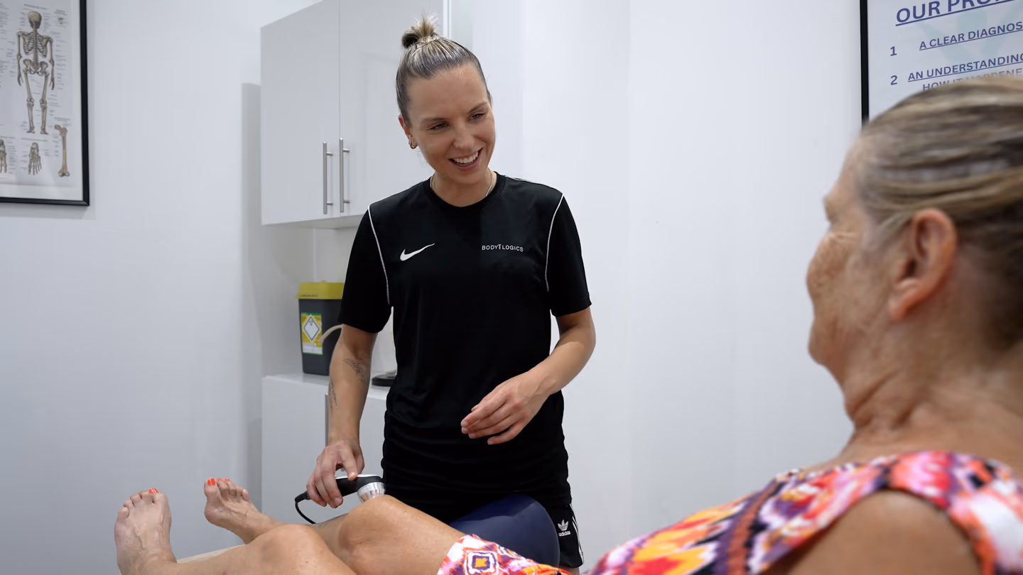 A Bodylogics clinician smiling while treating an older patient's knee with a handheld therapy device in a clinic treatment room